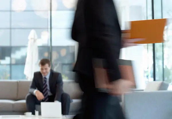 Eine unscharfe Silhouette im Anzug läuft von links nach rechts. Dahinter sitzt ein Mann auf einem Sofa.